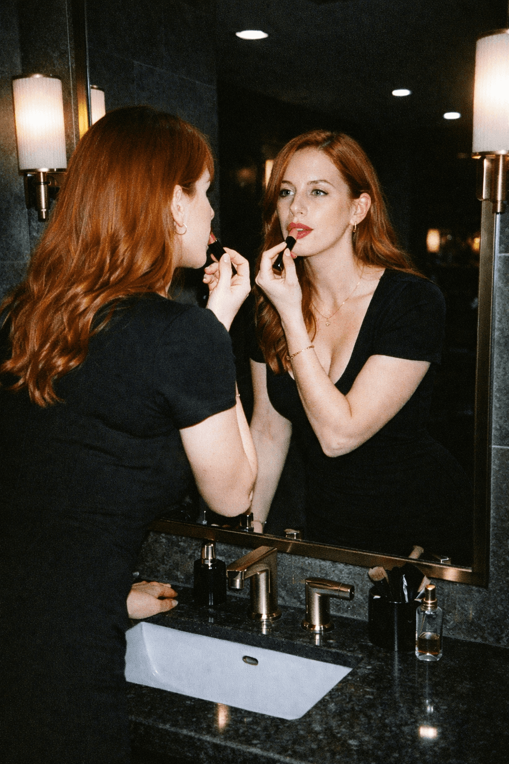 Woman applying lipstick in a mirror inside a dark bathroom, lit by a harsh direct flash that brightens her face while the background falls into shadow.