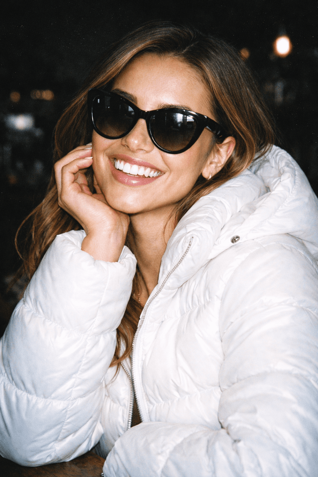 Close-up portrait of a smiling woman wearing black sunglasses and a white puffer jacket, lit by a harsh direct flash that brightens her face while the background falls into dark shadows with subtle grain.
